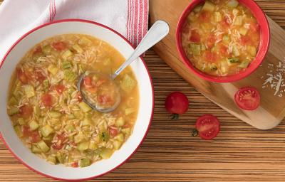 Fotografia em tons de branco, vermelho e marrom, sopeira branca com borda vermelha contendo sopa com legumes, arroz e uma concha, ladeada por guardanapo branco e vermelho, ao lado pote com sopa sobre tábua, tomate, tudo sobre bancada em tons de marrom.