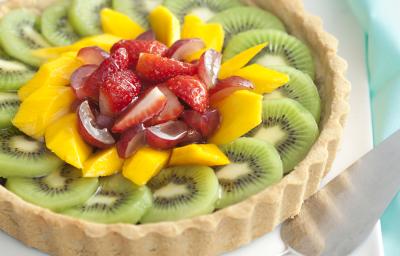 Fotografia em tons de verde, branco, amarelo e vermelho, com torta de frutas sobre porcelana branca com uma espátula e guardanapo verde ao lado, tudo sobre bancada em tons de branco e cinza.