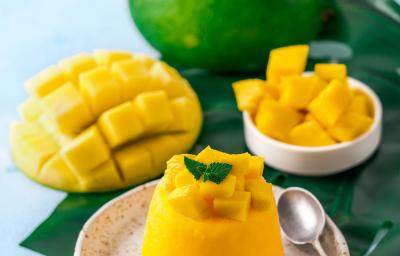 Fotografia de um flan de manga e calda de carambola, com pequenos pedaços de manga por cima. Ao lado da sobremesa, uma colher pequena que está sobre um miniprato de cerâmica branco com pontos pretos. Ao fundo, manga em pedaços e uma inteira.