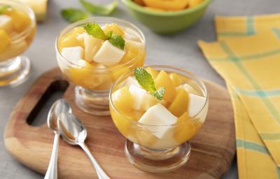 Foto da receita de Gelatina de Verão. Observa-se três potes individuais de vidro com as gelatina em quadrados e frutas amarelas picadas sobre uma tábua de madeira. Ao lado dos potes, duas colherinhas acompanham.