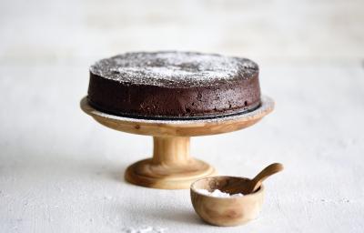 Foto da receita de Bolo Chocolatudo Sem Farinha. Observa-se uma boleira de madeira em um fundo branco com o bolo em cima, polvilhado de açúcar.