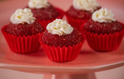 Fotografia de cinco unidades forminhas de cor vermelha, um junto da outra, e dentro de cada forminha tem um brigadeiro de red velvet, passado no açúcar cristal com uma decoração de cream cheese. Os brigadeiros estão sobre um apoio decorativo rosa alto.