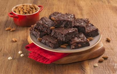 Fotografia em tons de marrom e vermelho de uma bancada de madeira com uma tábua de madeira e um paninho vemelho, sobre ele um prato redondo com pedaços de brownie. Ao fundo um recipiente redondo com amêndoas.