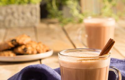 Fotografia em tons de marrom em uma bancada de madeira com um pano azul e duas xícaras de vidro com o chocaccino e um pau de canela para enfeitar. Ao fundo, um prato com cookies