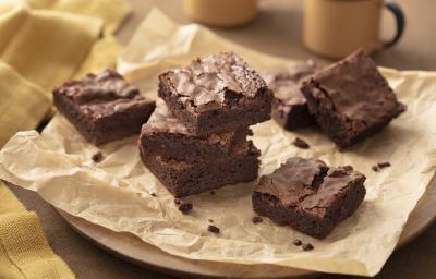 Fotografia em tons de marrom e bege em uma bancada de madeira, um pano bege, uma tábua de madeira redonda, um papel manteiga marrom com vários brownies de chocolate em cima. Ao fundo, xícaras beges.