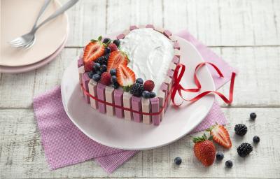 fotografia em tons de branco e rosa de uma bancada branca vista de cima, ao centro um pano rosa com um prato redondo por cima um bolo em formato de coração com marshmallow, morangos e frutas vermelhas por cima para decorar.