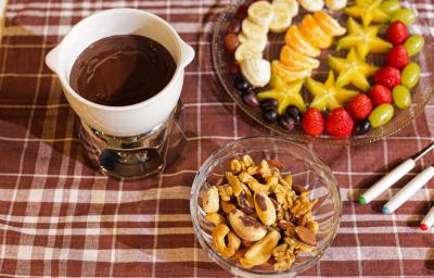 Fotografia em tons de marrom em uma bancada com uma toalha xadrez em marrom, com uma pequena panela branca com o fondue de chocolate dentro, ao fundo um recipiente com frutas picadas e um potinho de vidro com castanhas e amêndoas.