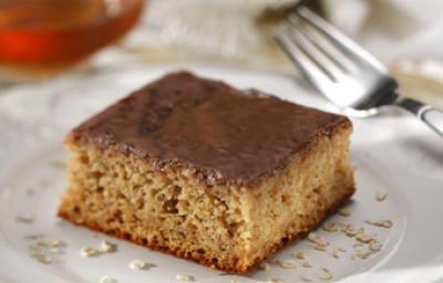 Fotografia em tons de marrom em uma mesa com uma toalha branca e bege, um prato branco raso redondo com uma fatia do bolo de banana com aveia e mel. Ao fundo, um potinho de mel.