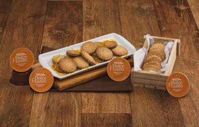 Fotografia em tons de marrom em uma bancada de madeira escura, um pano marrom com uma tábua de madeira em cima dele e um recipiente branco com vários biscoitos amanteigados com raspas de laranja em cima deles. Ao lado, uma caixinha com mais biscoitos.