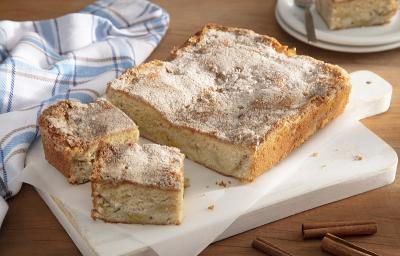 Foto em tons de bege da receita de cuca de banana tradicional servida sobre uma tábua branca em cima de uma mesa de madeira com um pano azul quadriculado ao lado além de alguns paus de canela em volta