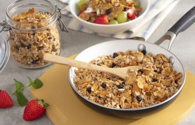 Fotografia em tons de amarelo em uma bancada de madeira com uma tábua de madeira, uma frigideira grande dentro com a granola com uvas-passas, cereal matinal dentro. Ao lado, um potinho com a granola e ao lado um outro potinho branco com frutas.