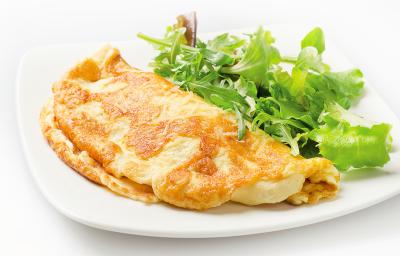 Fotografia de um omelete com crosta de queijo crocante dobrado ao meio, ao lado de uma salada de folhas, sobre um prato de vidro branco. O prato está sobre uma mesa branca.