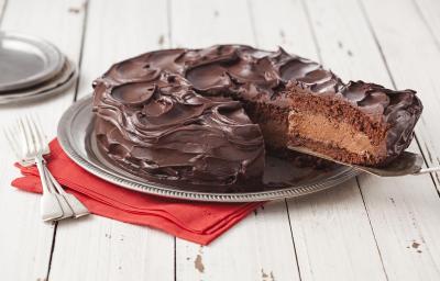 Fotografia em tons de branco e marrom de uma bancada de madeira, um paninho vermelho, um prato redondo prateado, dois garfos prateados e um bolo de chocolate com uma fatia sendo retirada, mostrando o recheio de mousse.