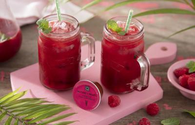 Foto em tons de cor de rosa da receita de pink lemonade com frutas vermelhas e hortelã servida em canecas altas de vidro em cima de uma tábua rosa clarinha com um pote de framboesas ao lado