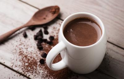 Fotografia em tons de branco e marrom tirada de uma bancada marrom, contém uma xicara branca com chocolate quente dentro e ao lado uma colher de madeira e chocolate em pó espalhado pela bancada.