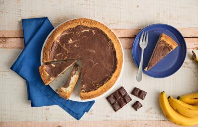 Fotografia em tons de azul em uma bancada de madeira, um pano azul, um prato grande raso com a cheesecake de banana com chocolate em cima dele. Ao lado, um prato pequeno azul raso com uma fatia da cheesecake, um cacho de banana e quadradinhos de chocolate