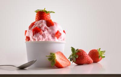 Fotografia de frozen iogurte de morango em um potinho de papel com pedaços de morango, e em cima, um morango inteiro. Ao lado, dois morangos inteiros e um morango cortado pela metade, no outro lado, uma colher de sobremesa, sobre um fundo branco.