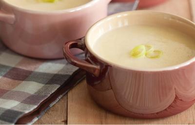 Fotografia em tons de rosa em uma bancada de madeira com uma toalha xadrez preta e rosa, duas mini panelinhas de cerâmica rosa com a sopa de alho-poró dentro delas.