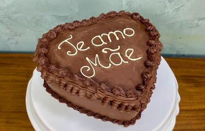 Fotografia em tons de castanho com um prato branco ao centro. Em cima do prato existe um bolo alto em formato de coração com cobertura de chocolate. Em cores brancas existe um escrito "Te amo Mãe"