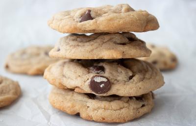 Fotografia de quatro cookies de leite condensado e baunilha empilhados, todos têm gostas de chocolate. Os cookies estão sobre uma mesa branca, ao fundo, tem mais unidades deles, espalhados.