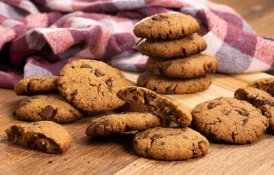 fotografia em tons de marrom, vinho e roxo de uma bancada marrom vista de frente, contém diversos biscoitos de chocolate espalhados pela mesa, e ao fundo  um pano xadrez em tons de vinho e roxo.