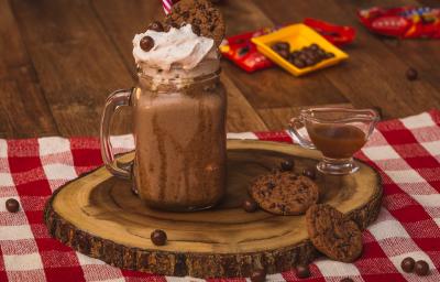 Foto de uma bancada de madeira. Sobre ela há uma toalha quadriculada vermelho e branco, uma tábua de madeira com um copo com o Milkshake de Nescau decorado com um canudo vermelho. Ao lado há um potinho com calda, 2 cookies e algunmas bolinhas de chocolate