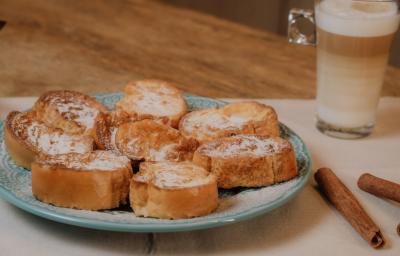 Fotografia em tons de marrom de uma bancada de madeira com um paninho branco sobre ele um prato azul claro e rabanas com açúcar e canela. Ao fundo paus de canela com um copo de leite.