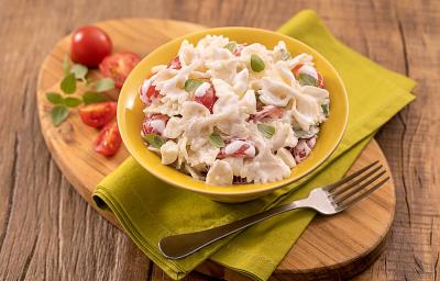 Foto da receita de saladinha de macarrão no copinho servida em um copo largo amarelo sobre um paninho verde com um garfo prateado ao lado, há também, em cima da tábua de madeira, tomates cerejas cortados ao meio