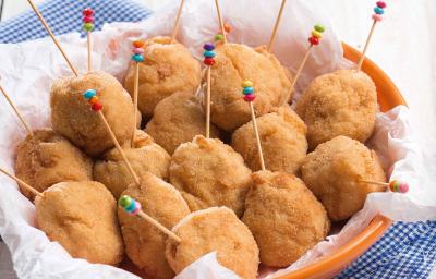 Fotografia em tons de azul em uma bancada de madeira clara com um pano azul quadriculado e em cima um prato redondo fundo laranja com coxinhas cremosas de frango dentro dele e palitinhos coloridos espetados nelas.