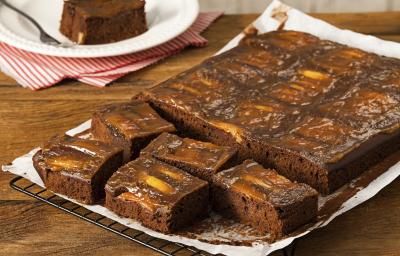 fotografia em tons de marrom e branco de uma bancada marrom vista de frente. Contém uma grade com um papel branco por cima, que contém um bolo de chocolate com pedaços de maçãs e cortado em quadrados