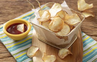 Fotografia em tons de amarelo e verde em uma bancada de madeira, com um pano listrado em amarelo e verde e uma tábua de madeira com uma cestinha de tela com várias batatas chips. Ao lado, um potinho amarelo com ketchup.