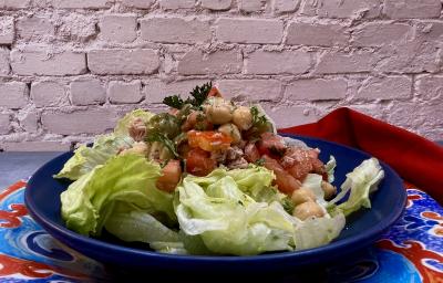 Foto da receita de Salada de Grão de Bico. Observa-se um prato azul redondo fundo com a salada com tomate, grão de bico, atum e alface americana dentro. Ao fundo, uma parede de tijolo branco.