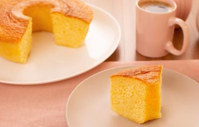 Fotografia de um prato raso branco grande com um bolo quase inteiro de fubá, na frente tem outro prato, menor e de tom mais escuro, com uma fatia do bolo de fubá. Um pouco mais ao fundo tem uma xícara rosa com café.