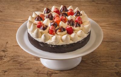 Foto aproximada de uma torta num prato branco alto. A torta tem uma base de biscoito escura e está decorada com chantilly, suspiros pequenos e morango, alguns banhados em chocolate. Tudo está numa bancada de madeira.