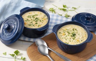 Fotografia em tons de branco, azul e marrom de uma bancada de madeira branca com um tábua de madeira, sobre ela um recipiente redondo azul com a sopa e duas colheres. Ao lado um paninho xadrez branco e azul com um recipiente azul com a sopa.