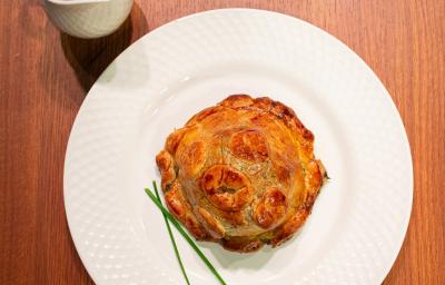 Fotografia de uma receita de pithivier paraense com duxelles de ora-pro-nóbis sobre um prato raso branco, o qual está sobre uma bancada de madeira. Ao lado do prato tem um recipiente fundo e pequeno com molho.