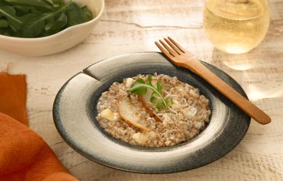 Foto da receita de risoto de pera com brie servida em um prato fundo azul com um garfo de madeira na borda e um copo de vidro com vinho branco ao fundo