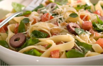 fotografia em tons de branco, verde e amarelo tirada de um recipiente redondo branco com macarrão, rúcula e pedaços de azeitona