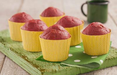 Fotografia em tons de verde em uma bancada de madeira clara, uma tábua de madeira verde, um paninho verde com bolinhas brancas e forminhas amarelas de muffin com o bolinho de beterraba dentro delas. Ao fundo, uma xícara verde