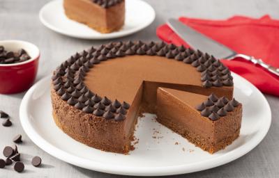 Fotografia em tons de branco e vermelho de uma bancada branca com um prato branco, sobre ele uma torta de chocolate. Ao lado um paninho vermelho com uma espátula. Ao fundo um prato branco com uma fatia de torta e um potinho com gotas de chocolate.