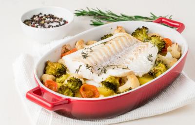 fotografia em tons de branco, vermelho e amarelo de uma bancada branca vista de cima. Contém um recipiente oval vermelho, com um pedaço de bacalhau e legumes em volta.