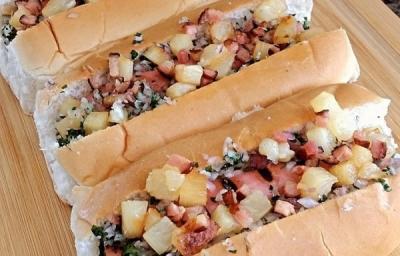 Foto em tons marrom claro da receita de cachorro quente havaiano servida em três porções uma ao lado da outra sobre uma tábua de madeira em cima de uma mesa de mármore