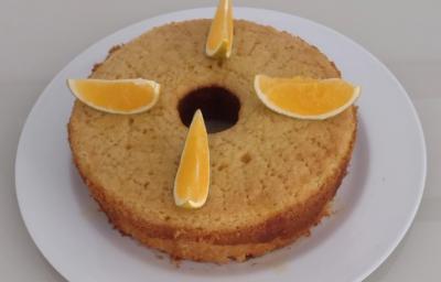 Fotografia de um bolo de laranja feito na forma de fundo central, sobre um prato de vidro branco. Sobre o bolo tem quatro pedaços de laranja cortados.