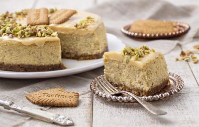 Foto da receita de Cheesecake de Pistache. Observa-se a torta com um pedaço cortado em um prato de sobremesa, decorado com pistaches e uma calda na cor branca.