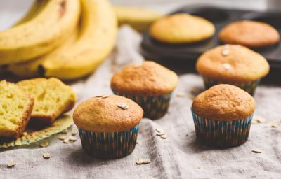 Fotografia de quatro muffins de banana e aveia dentro de uma forminha de papel colorida sobre uma toalha de mesa em tons de bege. Ao lado, um cacho de bananas, duas fatias do bolo, e ao fundo, dois muffins em uma forma preta de bolinhos.