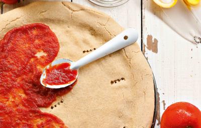 Fotografia em tons de vermelho e branco, de uma bancada de madeira com uma massa redonda de pizza, com molho de tomate pela metade e uma colher prateada. Ao redor potinhos, ovos, tomates e azeite.