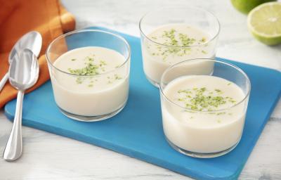 Foto da receita de mousse de limão zero lactose servida em três porções em taças sobre uma tábua azul com um pano laranja ao lado além de duas colheres prateadas