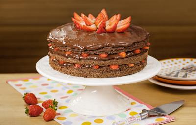 Fotografia em tons de marrom e vermelho de uma bancada de madeira, uma boleira com o bolo de chocolate e morangos. Ao lado quatro morangos para decorar, um espátula para servir e três pratos coloridos em tons de laranja com garfos.