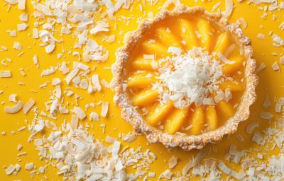 Fotografia vista de cima de uma torta tipo quiche com recheio cremoso de Leite MOÇA, suco de laranja e coco. A tortinha está sobre uma mesa amarela, e salpicada de pequenas lascas de coco.