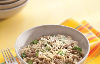 fotografia em tons de bege, cinza e amarelo de uma bancada bege vista de cima. Contém um pano amarelo com um recipiente redondo cinza com o risoto, com pedaços de carne e ao lado um garfo para servir.